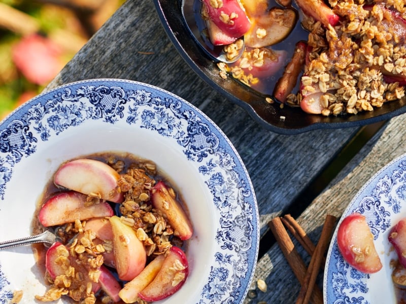 Bowl of apple crisp with cast iron skillet pictured at top of image