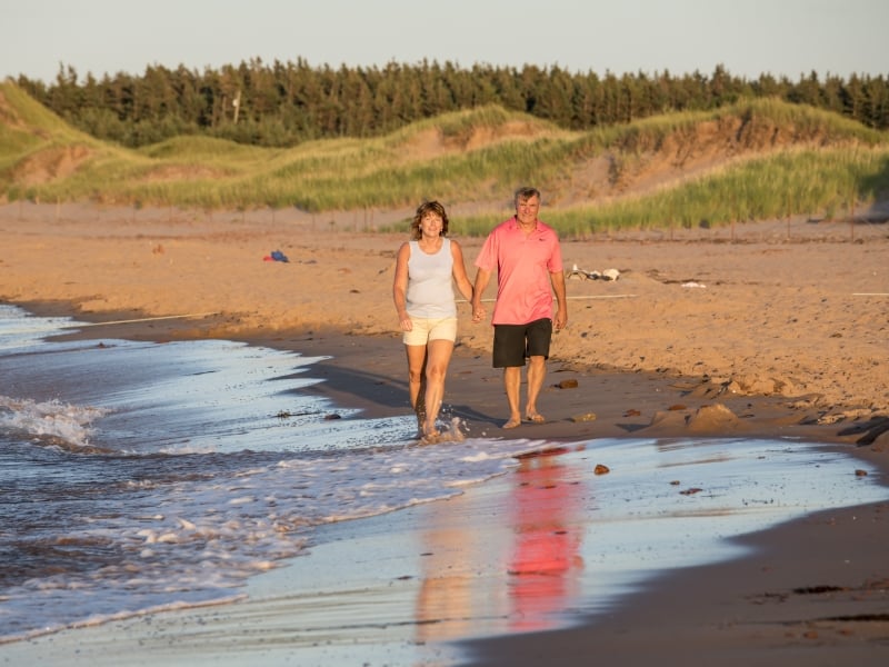 Couple walk at water's edge on Cavendish Beach with dunes in background