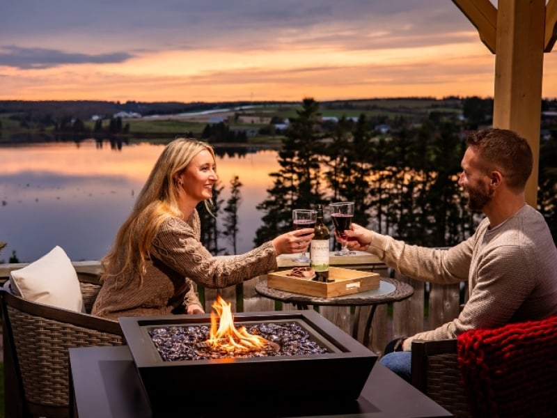 Couple sit on deck overlooking river at sunset with fireplace glowing