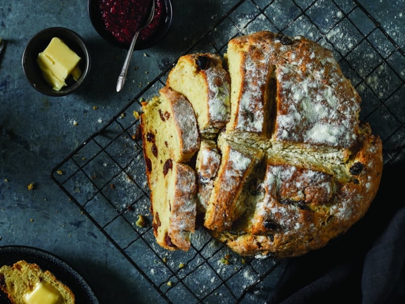 Overhead image of round loaf of potato soda bread on cooking rack with small bowls of butter and jam to the side