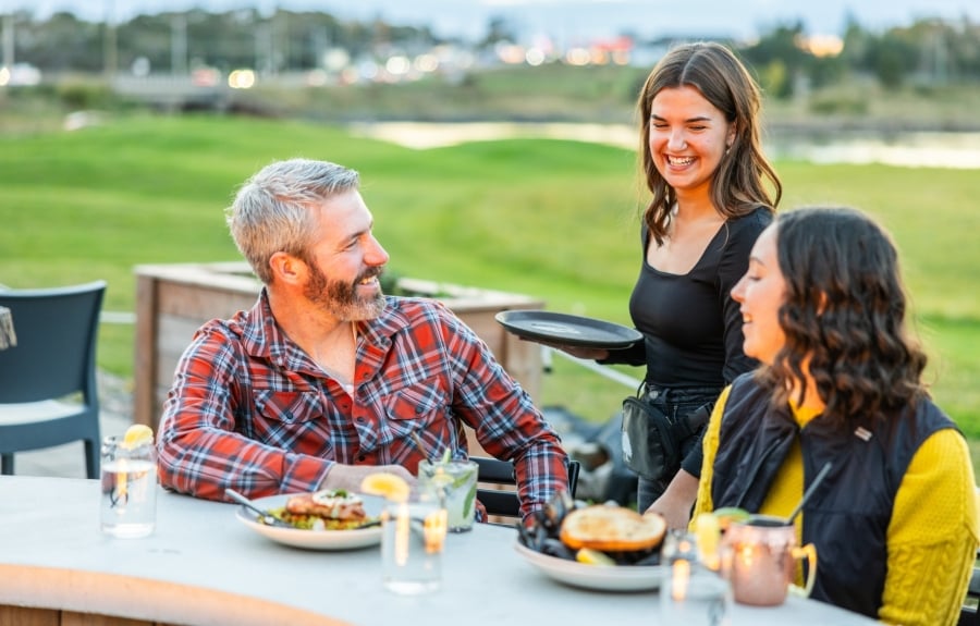 Two people dining on patio with server at the table with river in background