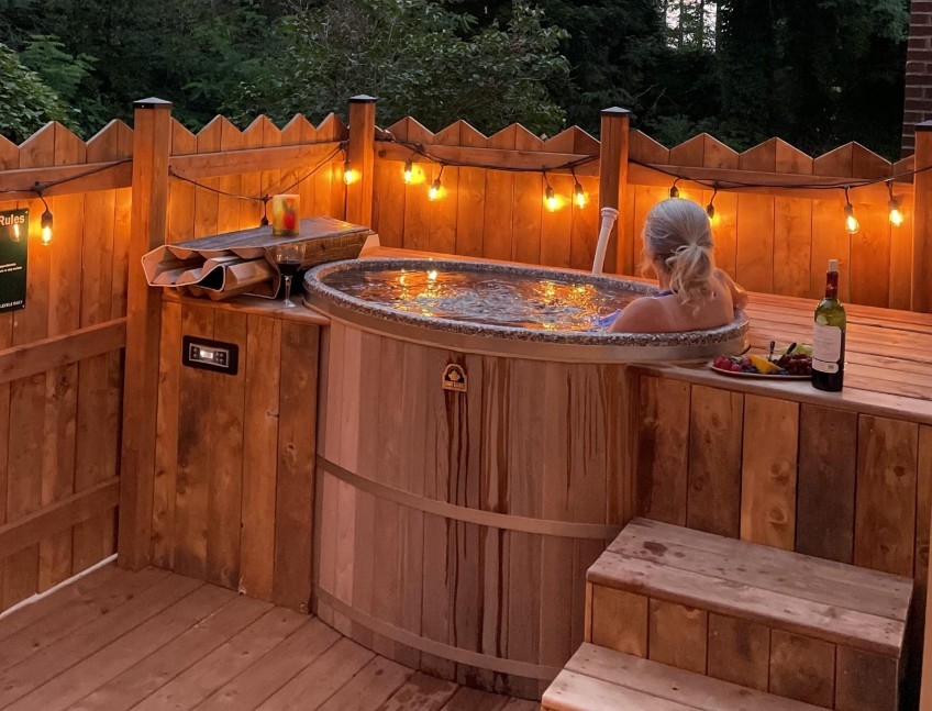 Female sitting in an outdoor cedar hot-tub surrounded by a wooden fence and outdoor lighting 