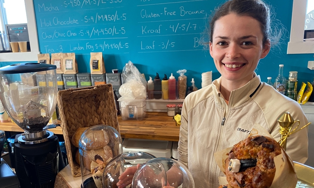 Female owner stands at front counter of Knead a Brake with menu on chalkboard on background