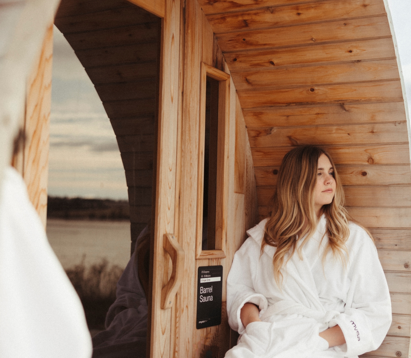 person sits outside barrel sauna at Mysa Nordic Spa overlooking river